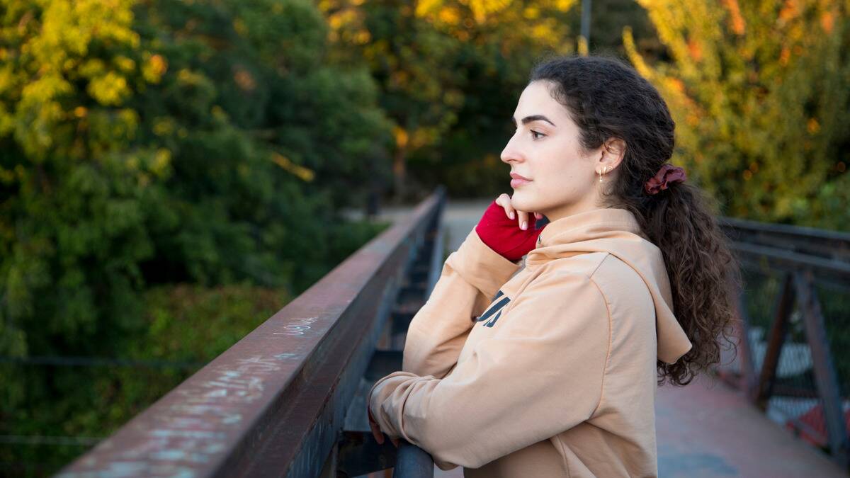 A woman standing on a small footpath bridge, leaning against the railing, looking out, a hand on her jaw.