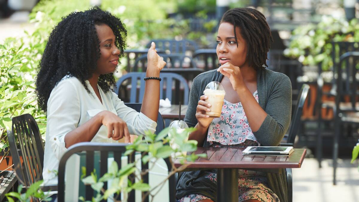 Two friends sitting at a cafe table, one talking and gesturing with her hands as the other sips her drink and listens.