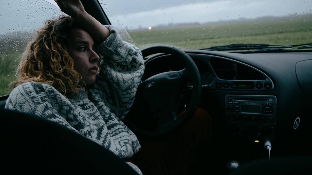 A woman sitting in her car, leaning against the driver's side door as it rains outside, her arm on her forehead, looking upset.