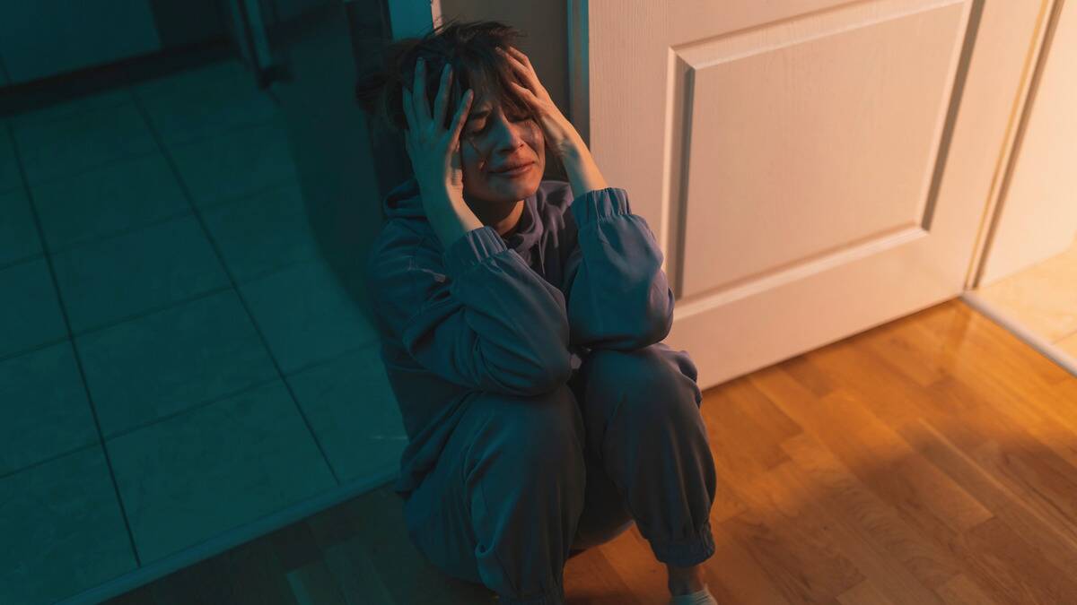 A woman sitting on the floor of her home, crying with her hands on the side of her head.