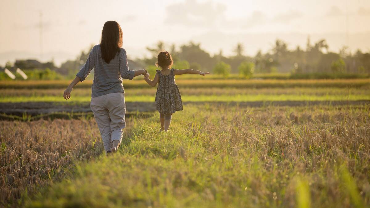 A mother holding hands with her young daughter as they walk outside, facing away from the camera, the daughter on a higher platform so she's even with her mom.