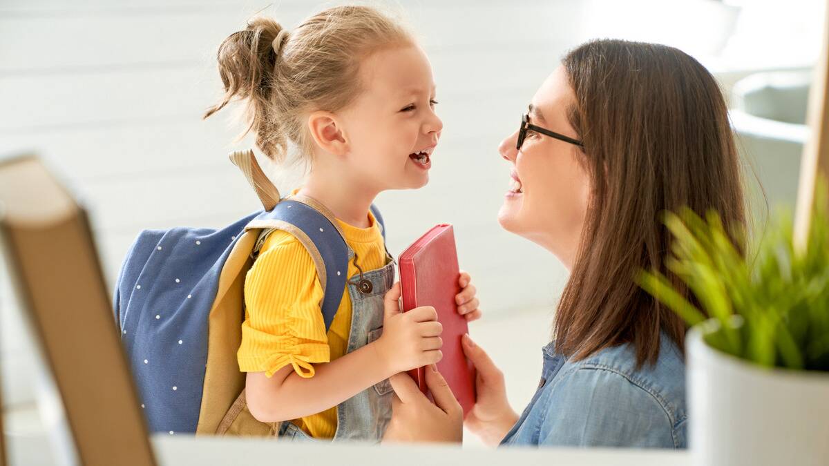 A woman kneeling down in front of her daughter as she hands her a book, both smiling excitedly.