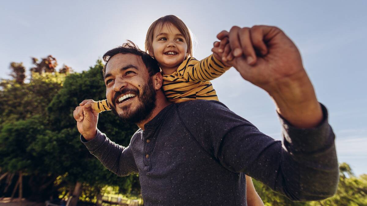 A low-angle shot of a man giving his child a piggyback ride, both of their arms outstretched.