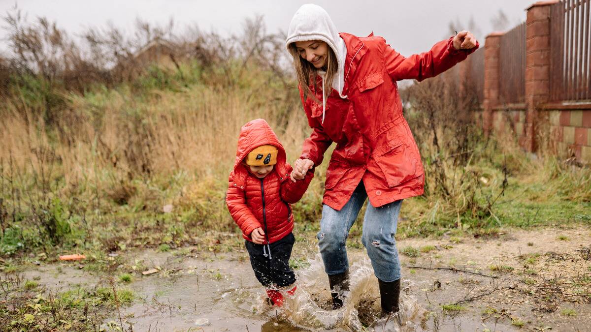 A mother holding hands with her young son as they both jump and splash in a puddle outside.