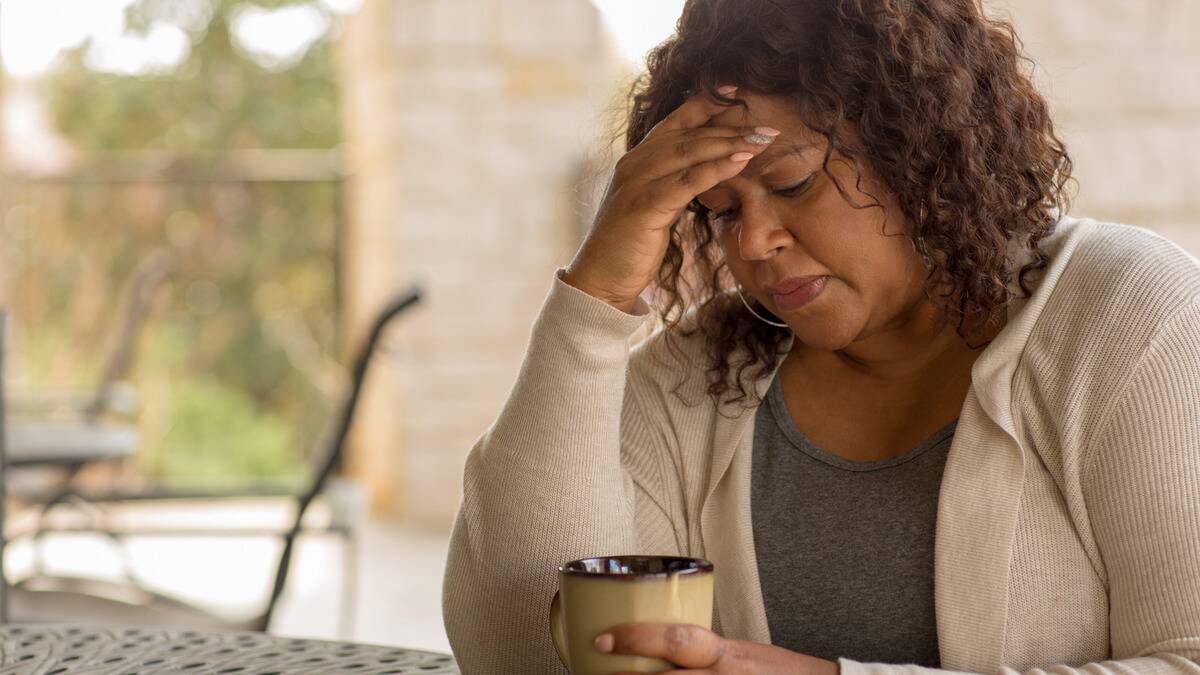 A woman sitting at a table, a mug in one hand, the other at her forehead.