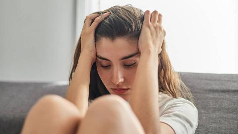 A close photo of a woman sitting on the ground, knees drawn up, hands in her hair.