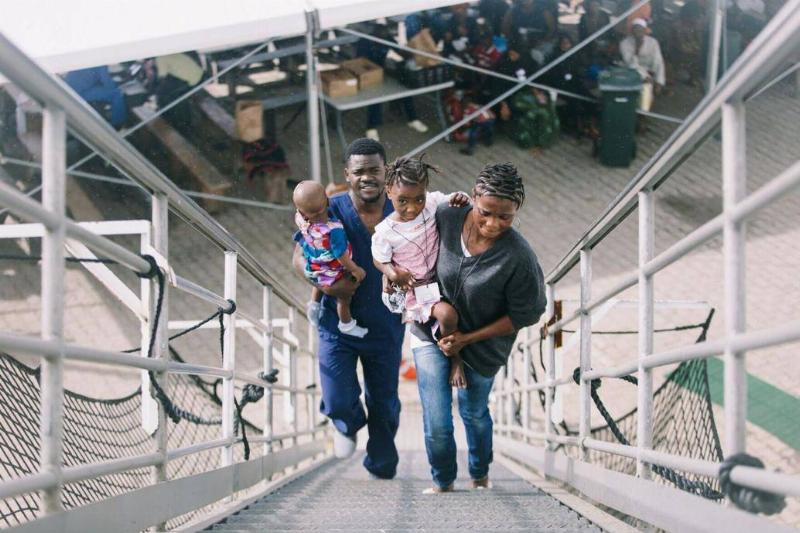 Gamai being carried by her mother up the steps to the ship, her father behind, carrying her younger sister.