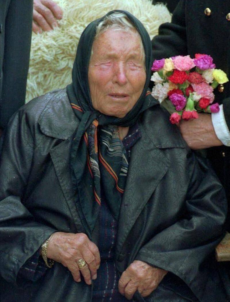 Baba Vanga sitting up, some people behind her, one holding a bouquet of flowers.