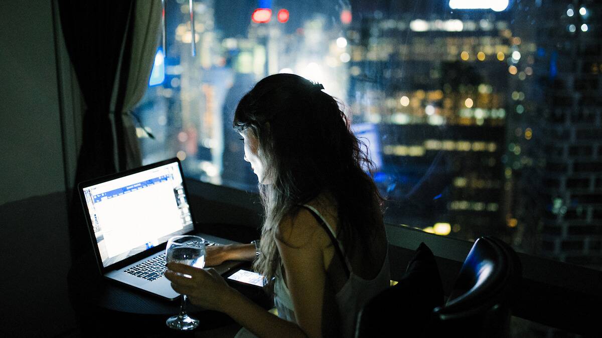 A woman working late at her laptop, the city visible outside her window.