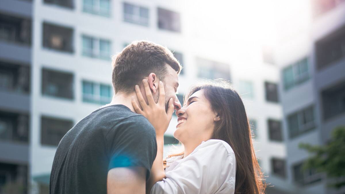 A photo of a couple standing outside, the woman holding her partner's face close to hers, both of them smiling.