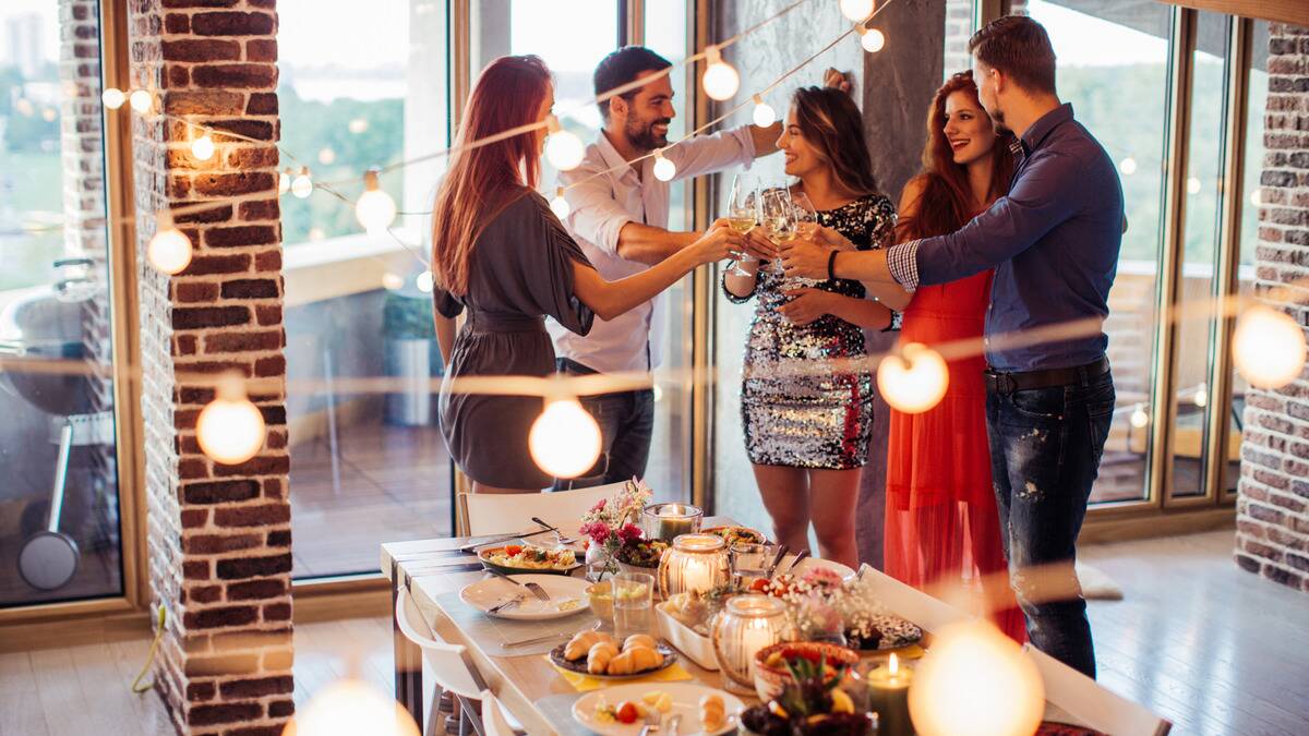 A group of friends standing, chatting, and cheersing their drinks together behind a table full of food.