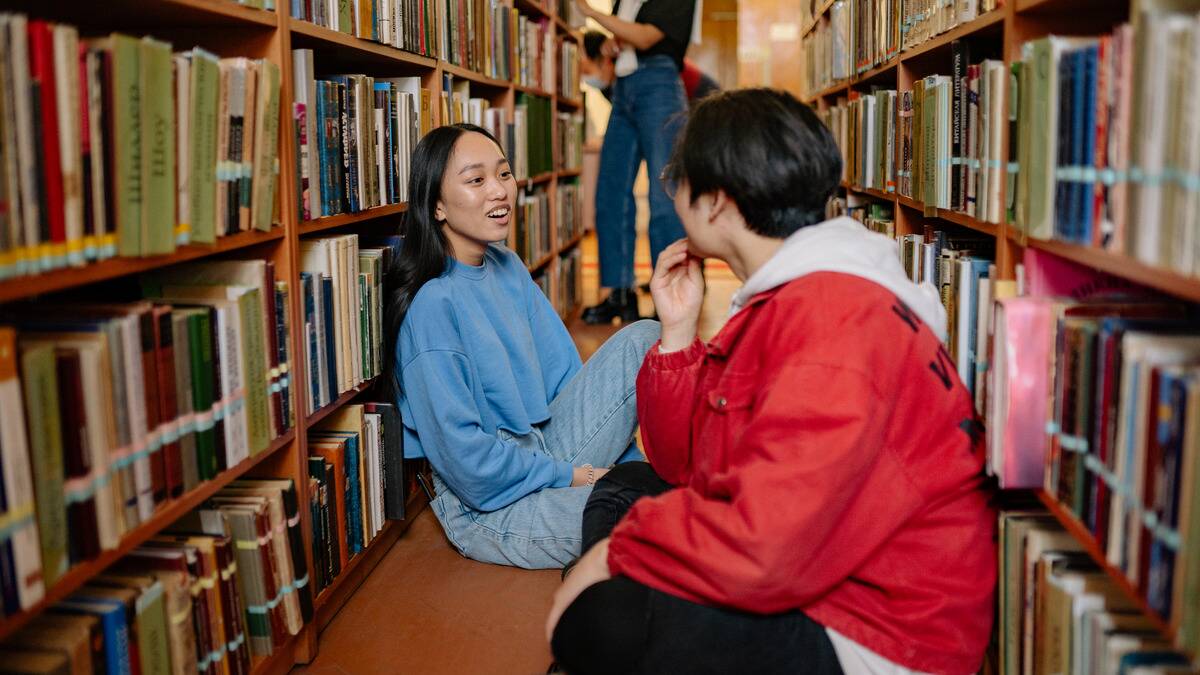 Two friends sitting on the floor in the aisle of a library, smiling and chatting.