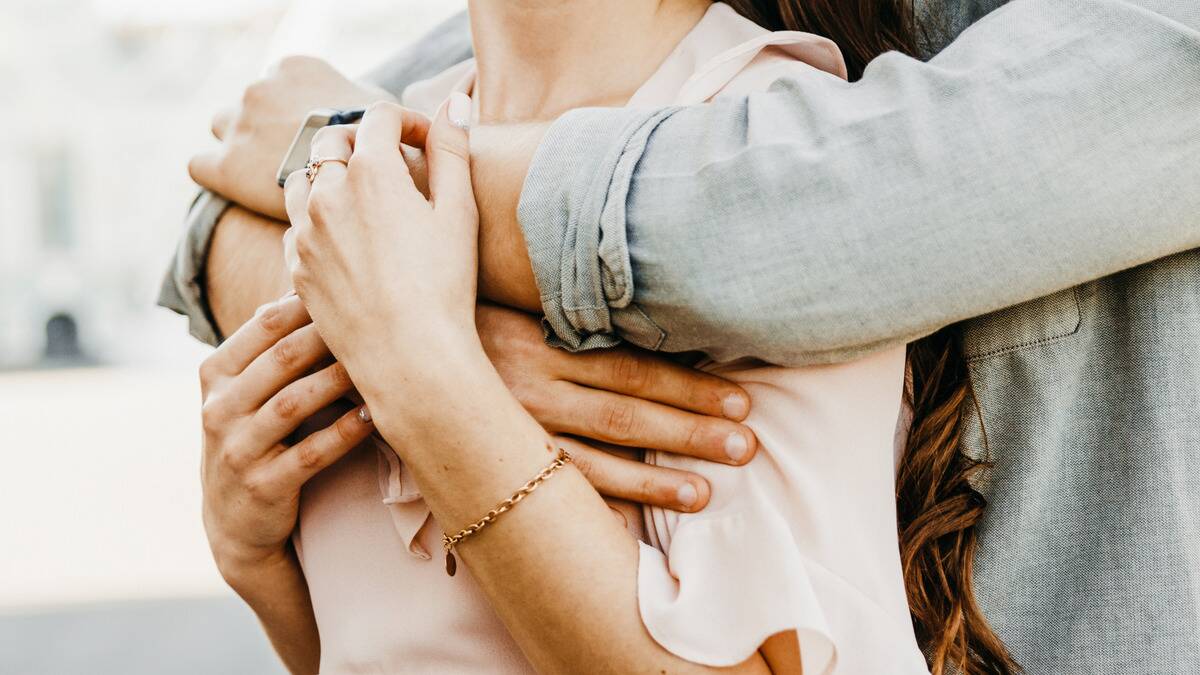 A torso photo of a woman who's being hugged from behind by her boyfriend, her hands coming up to touch his arms.