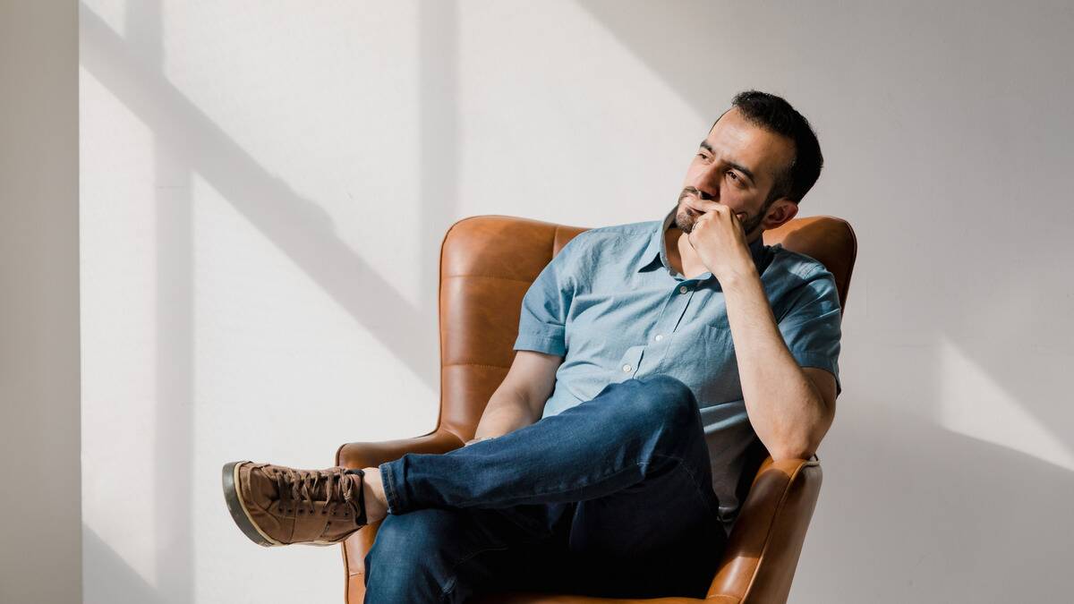 A man sitting in a chair against a blank wall, one leg folded over, a hand to his mouth, in deep thought.