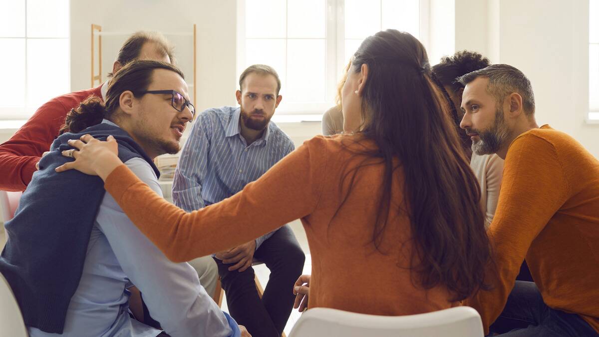 A support group session where one woman is reaching out to place a comforting hand on the shoulder of the man beside her.