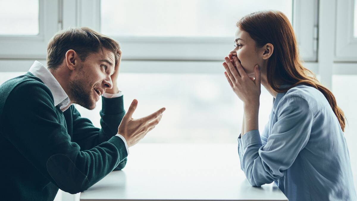 A couple sitting across a tabjle from one another, one appearing to be laughing with her hands covering her mouth, the other looking exasperated, a hand in his hair.