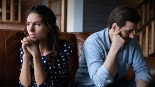 A couple sitting side by side on a couch but facing opposite directions, both looking sad and tired.