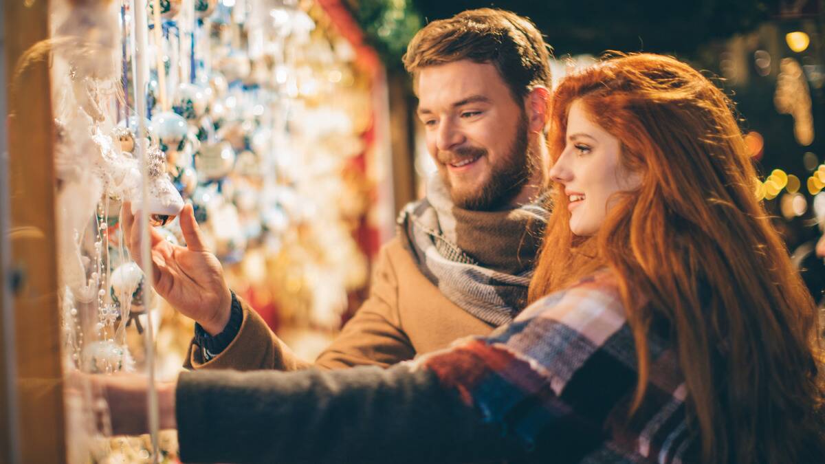 A couple at a Christmas market together, both reaching out to hold ornaments on display.