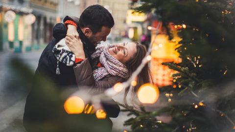 A couple on the street, by a Christmas tree and some lights, the man spinning the woman around as they both smile.