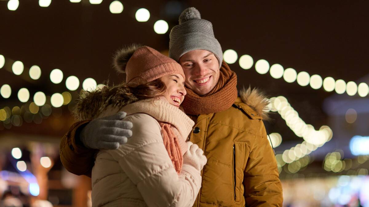 A couple in winter getup under some string lights, the woman leaning against the man's chest, the man with an arm around her, both smiling.