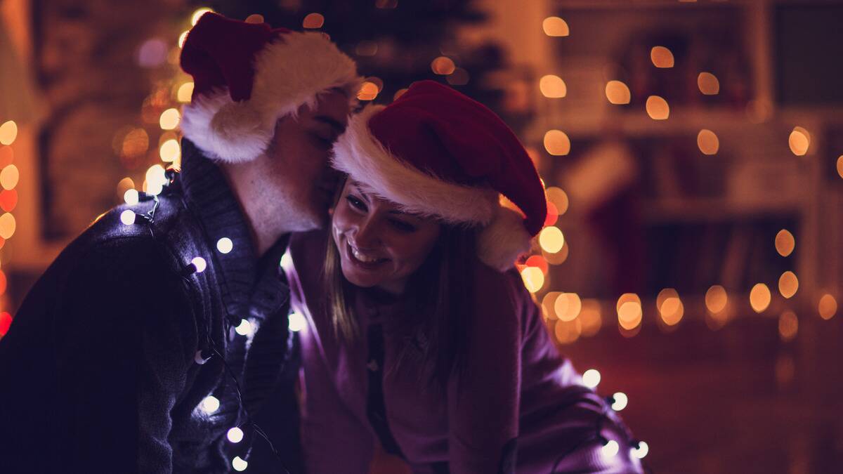 A couple, each wearing Santa hats, sitting wrapped in string lights, the man kissing the woman on the cheek as she smiles.