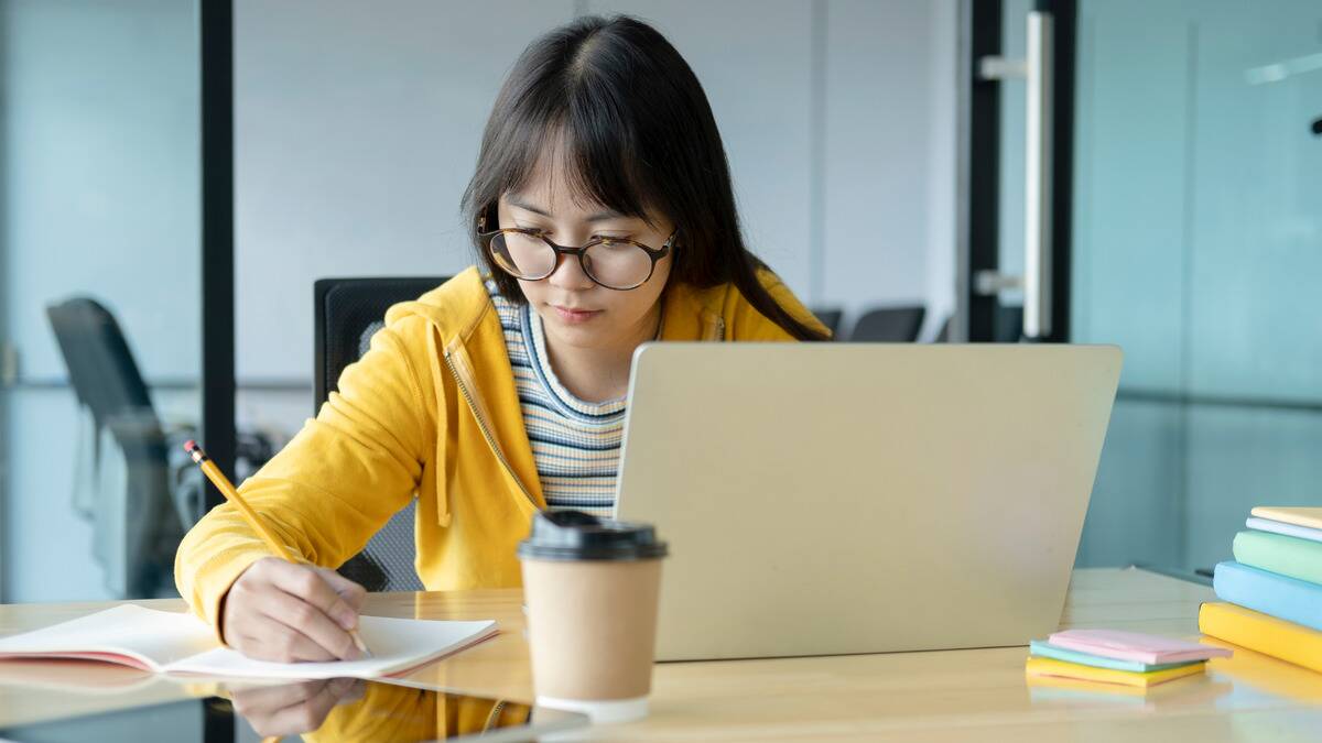 A woman sat at a table in front of her laptop, writing notes in a notebook at the same time.