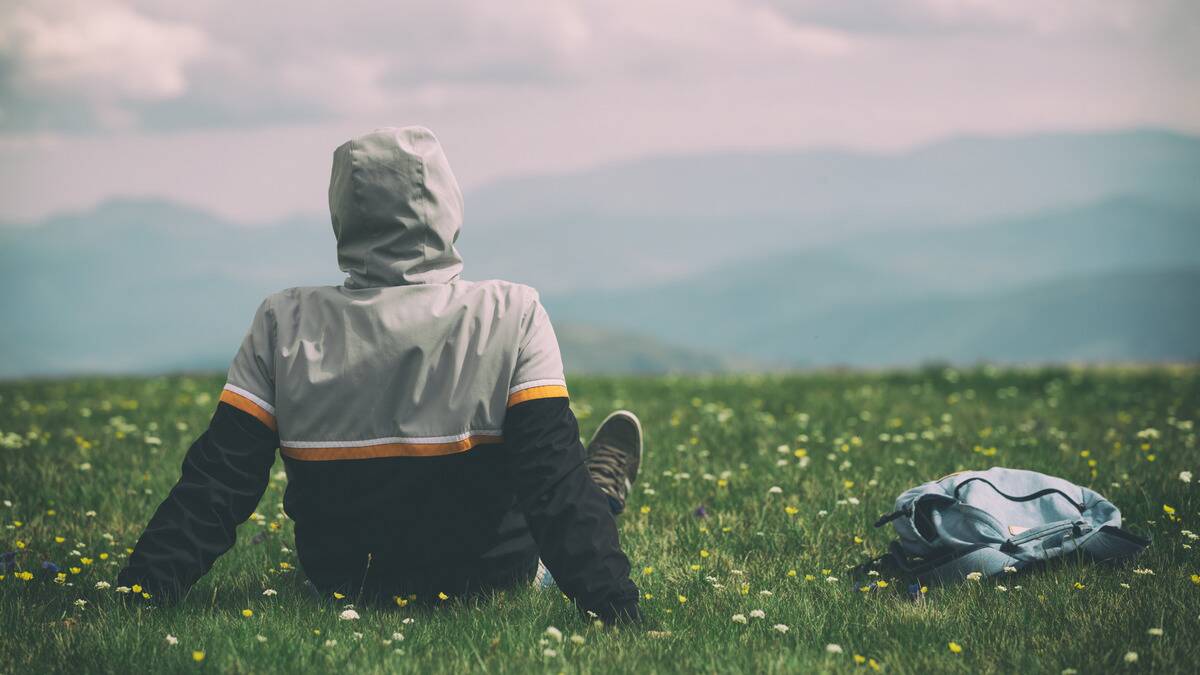 A person in a hooded raincoat sitting in a grassy field, facing away from the camera.