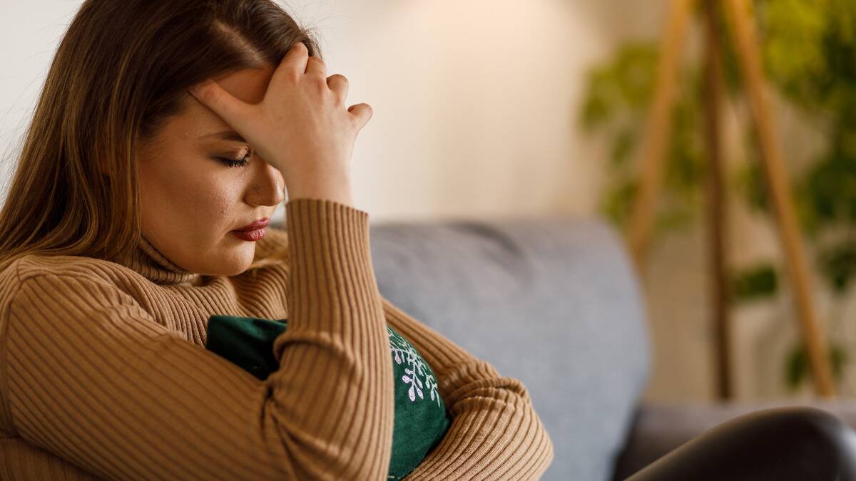 A woman sitting on her couch, a hand on her forehead, hugging a pillow, looking distressed.