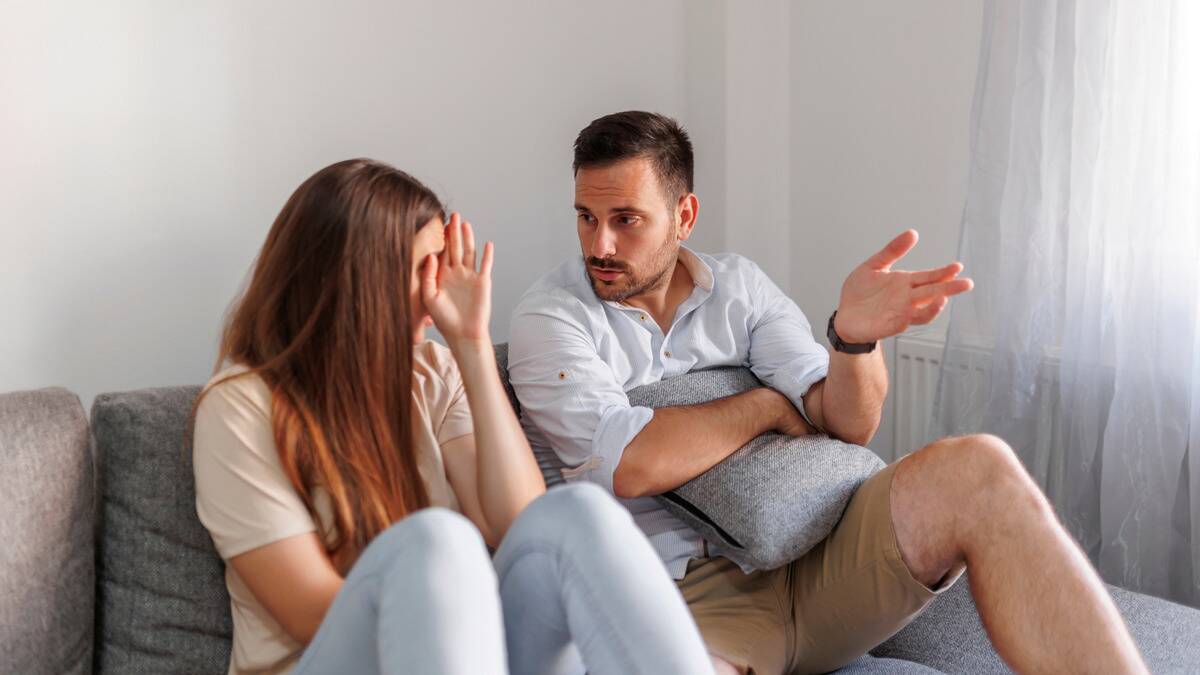 A man and woman sitting on a couch, the man talking and gesturing with his hand, the woman covering her face.