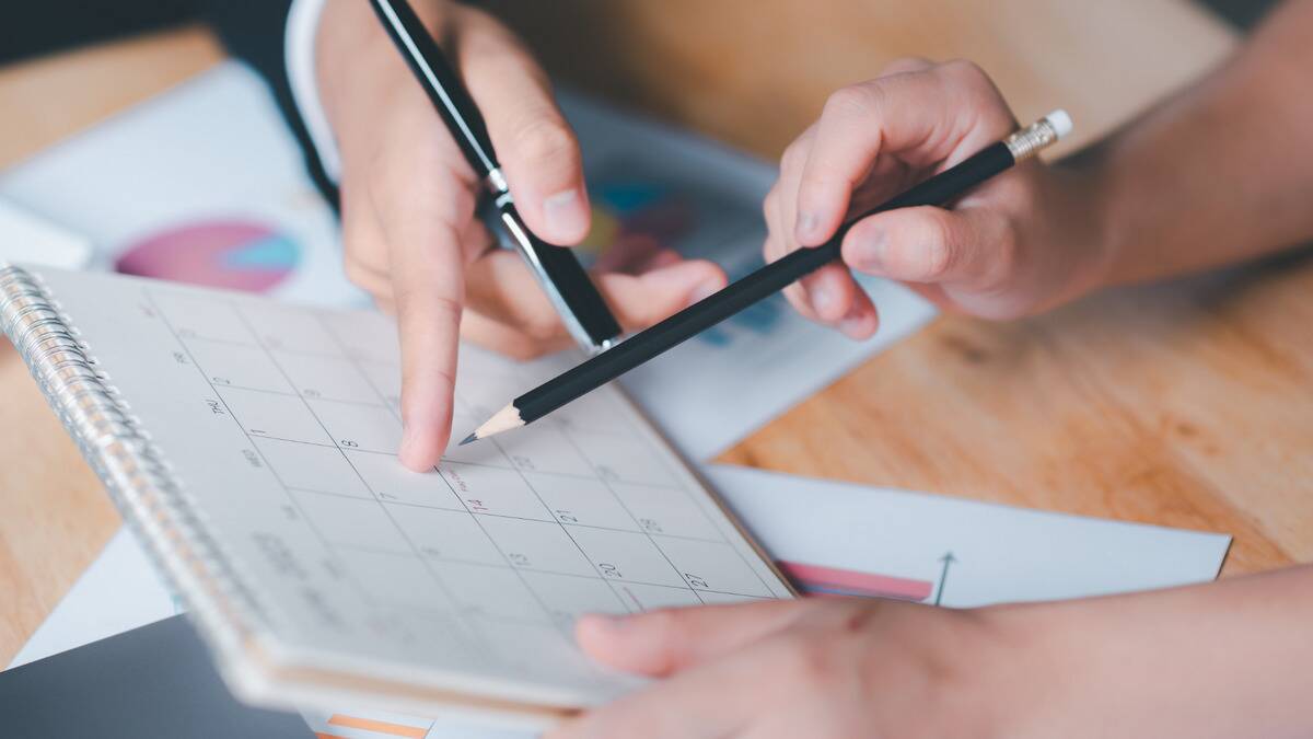 A close shot of a pair of hands, one holding a pen and one holding a pencil, pointing at a small monthly calendar.