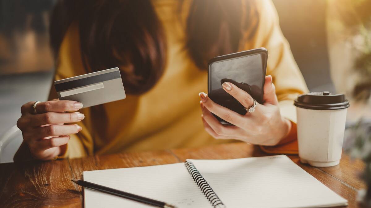 A woman sitting at a table holding her credit card in one hand and her phone in the other.