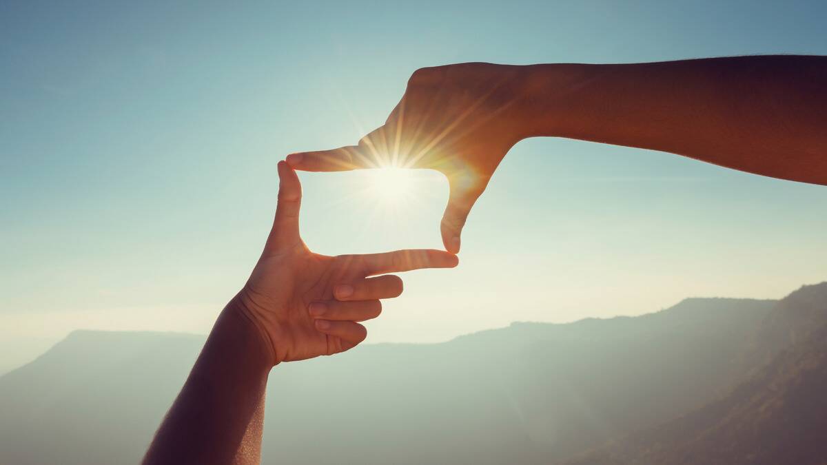 Two hands using their thumb and index fingers to make a rectangle shape, the sun shining through the box they made.