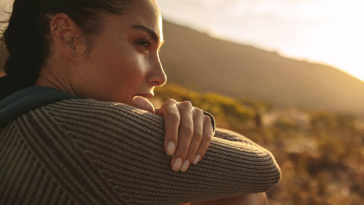 A close photo of a woman sitting outside at sunrise, arms folded atop her knees, her face tucked into them slightly.