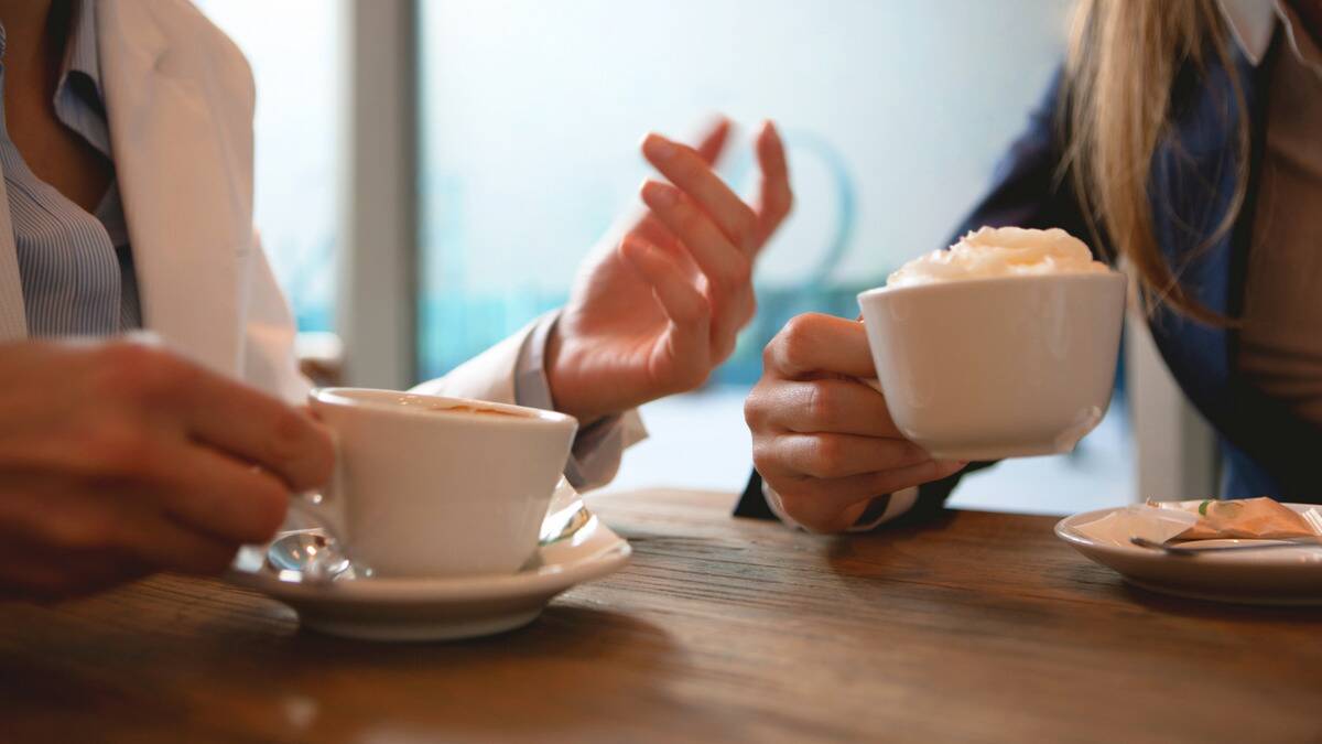 A close photo of two sets of hands at a cafe table, both holding a mug, one gesturing as its owner speaks.