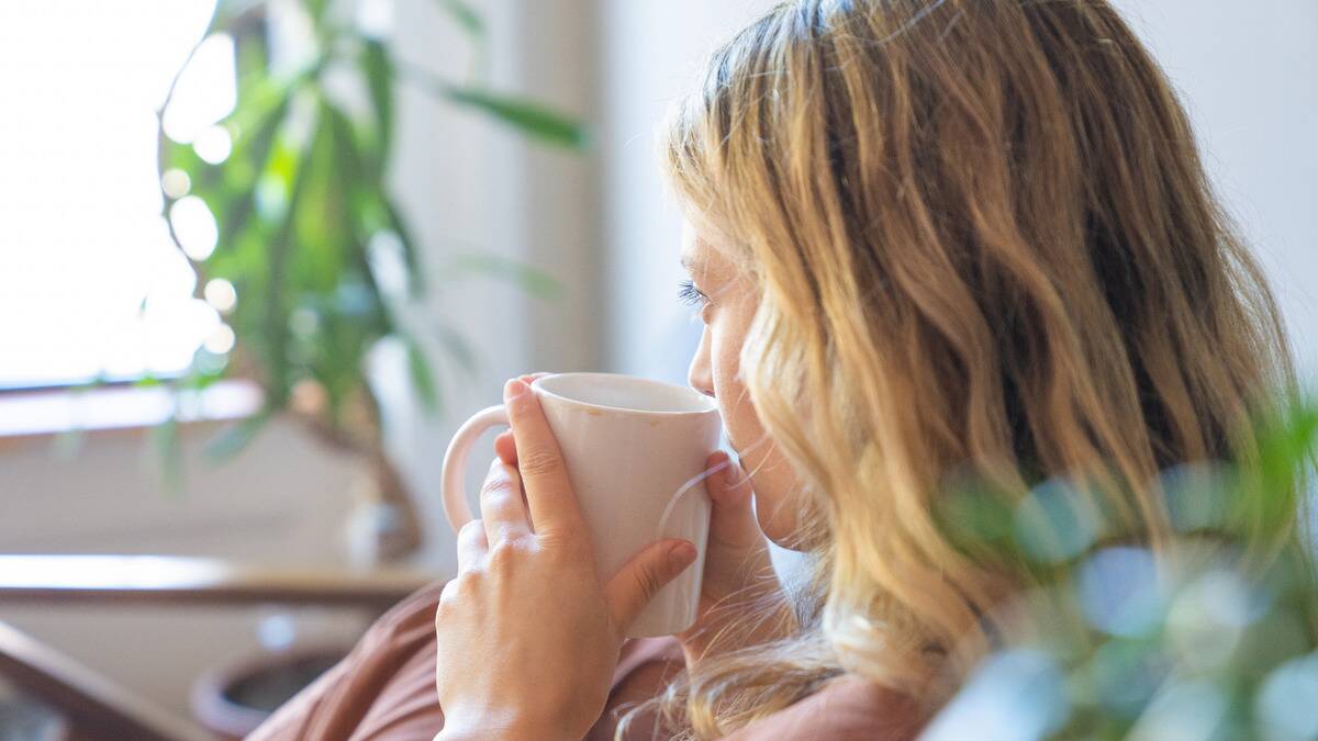 A photo from a slight behind angle of a woman sitting on her couch, holding a mug to her lips with both hands, looking forward toward the window.