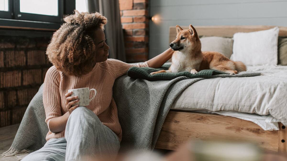 A woman sitting on her floor at the foot of her bed, a mug in one hand, using her other to pet a shiba inu that's on the bed.