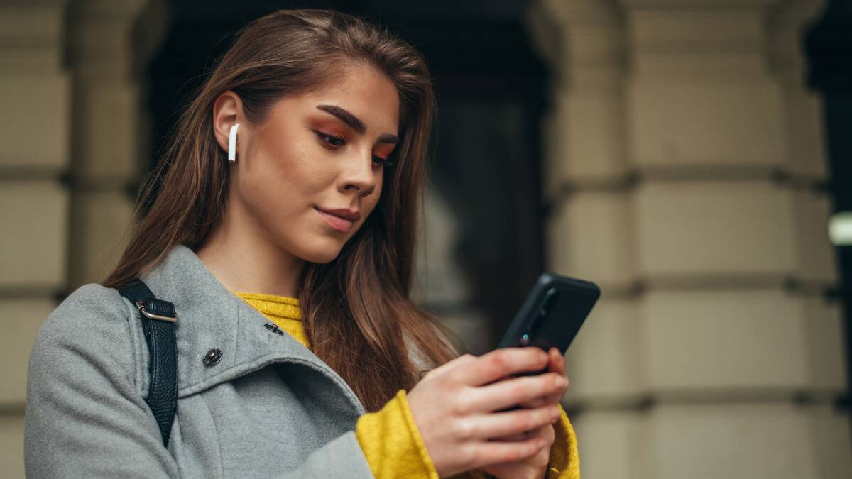 A young woman standing outside looking down at her phone, a slight smile on her face as she texts.