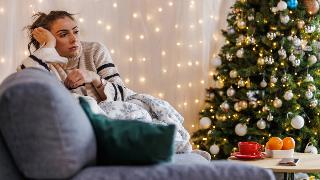 A woman sitting on a couch next to her Christmas tree, leaning her head into her hand, looking sad.