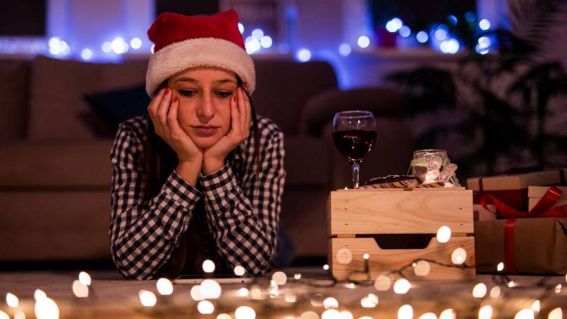 A woman laying on the floor in a Santa hat and some string lights on the ground, a wine glass on a box next to her, her chin in her hands.