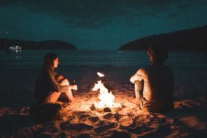 man and woman sit around bonfire