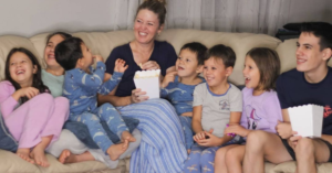 pam and 7 kids watching movie and laughing on couch with popcorn